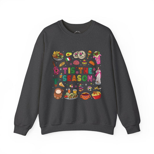 A dark gray crewneck sweatshirt featuring a colorful graphic print on the front with the text 'Tis the Season' surrounded by various cozy mexican illustrations like hot cocoa, burritos, tacos, pinata, and more seasonal traditions.