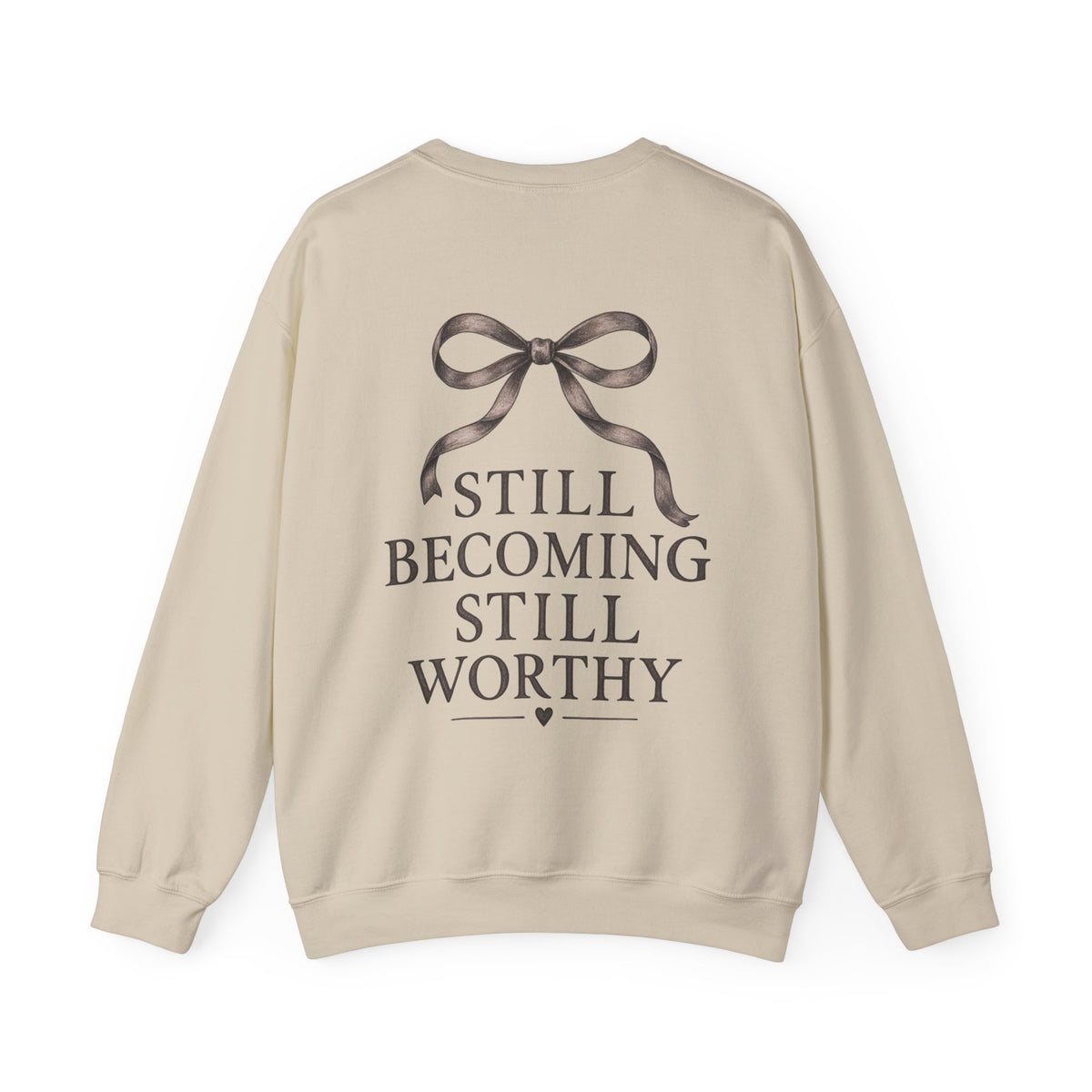 A beige crewneck sweatshirt shown from the back, featuring a dark brown graphic of a ribbon bow above the text 'STILL BECOMING STILL WORTHY' in a serif font.