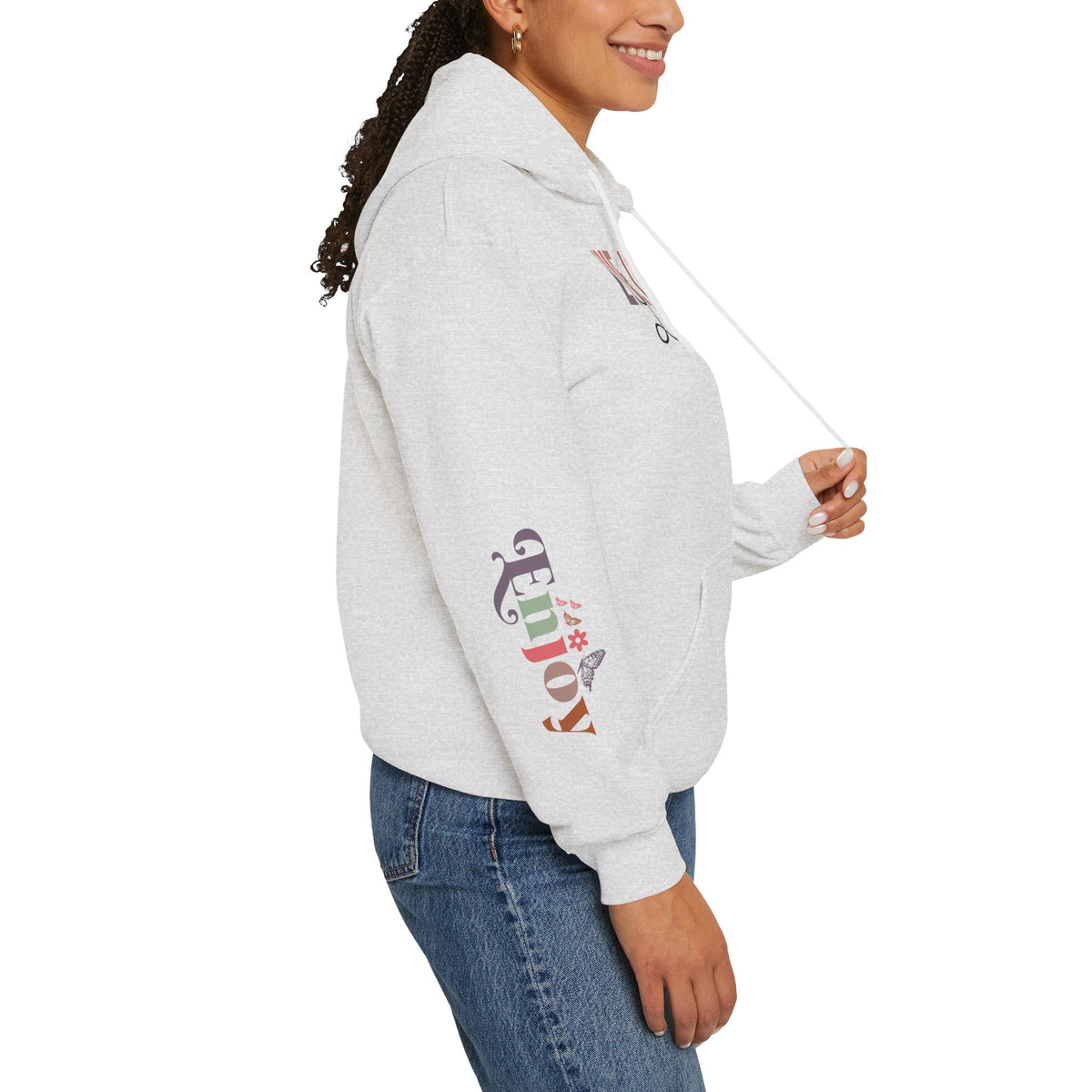 Person wearing a light heather gray hoodie with text graphics on the right sleeve 'ENJOY' with a small butterfly, paired with blue jeans, shown from a side profile against a white background.
