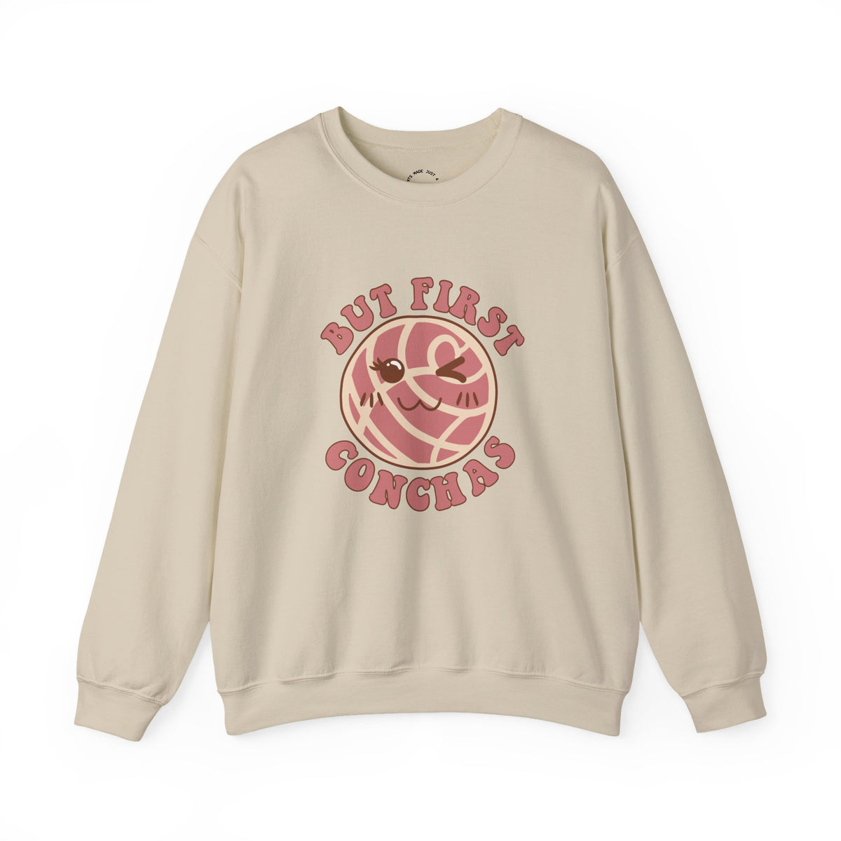 Beige crewneck sweatshirt featuring a graphic of a winking concha with the text 'BUT FIRST CONCHAS' in pinkish-red lettering.