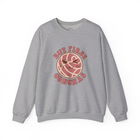 A heather gray crewneck sweatshirt featuring a graphic of a winking concha with the text 'BUT FIRST CONCHAS' in pinkish-red lettering.