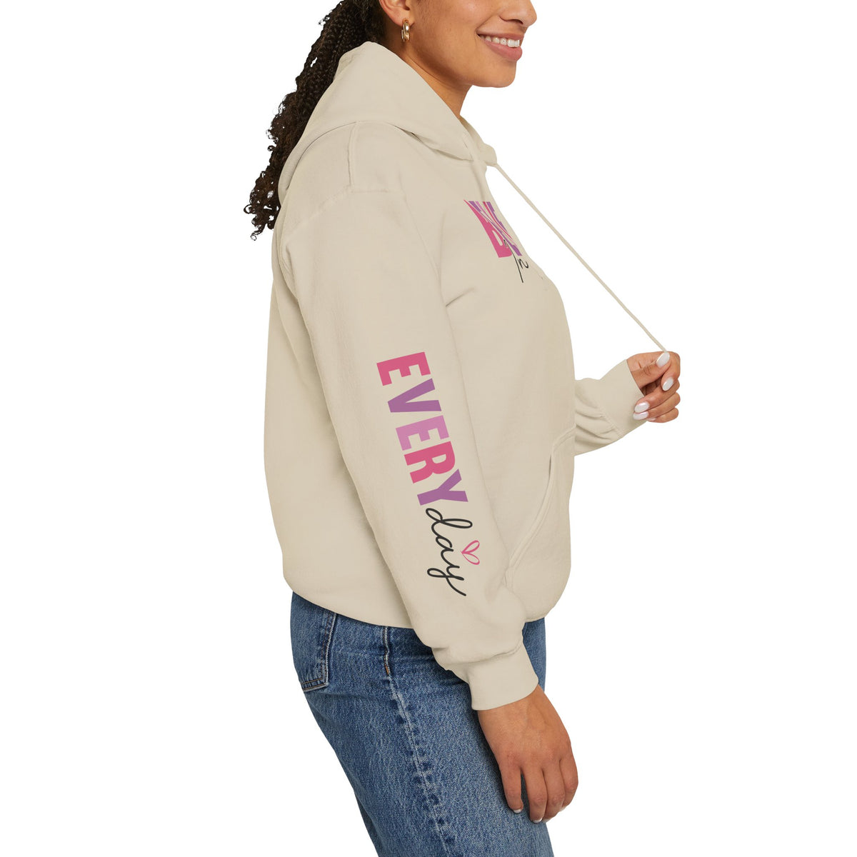 Person wearing a beige hoodie with pink and purple text graphics on the sleeve, paired with blue jeans, shown from a side profile against a white background.