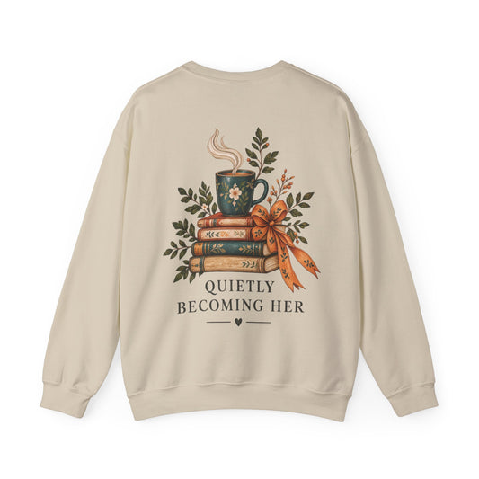 A beige crewneck sweatshirt shown from the back, featuring a graphic of a steaming mug on a stack of books tied with a ribbon, surrounded by green leaves, with the text 'QUIETLY BECOMING HER'.