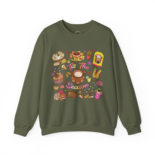 An olive green crewneck sweatshirt featuring a colorful Christmas-themed graphic print with illustrations of seasonal hispanic treats and the text 'Tis The Season'.