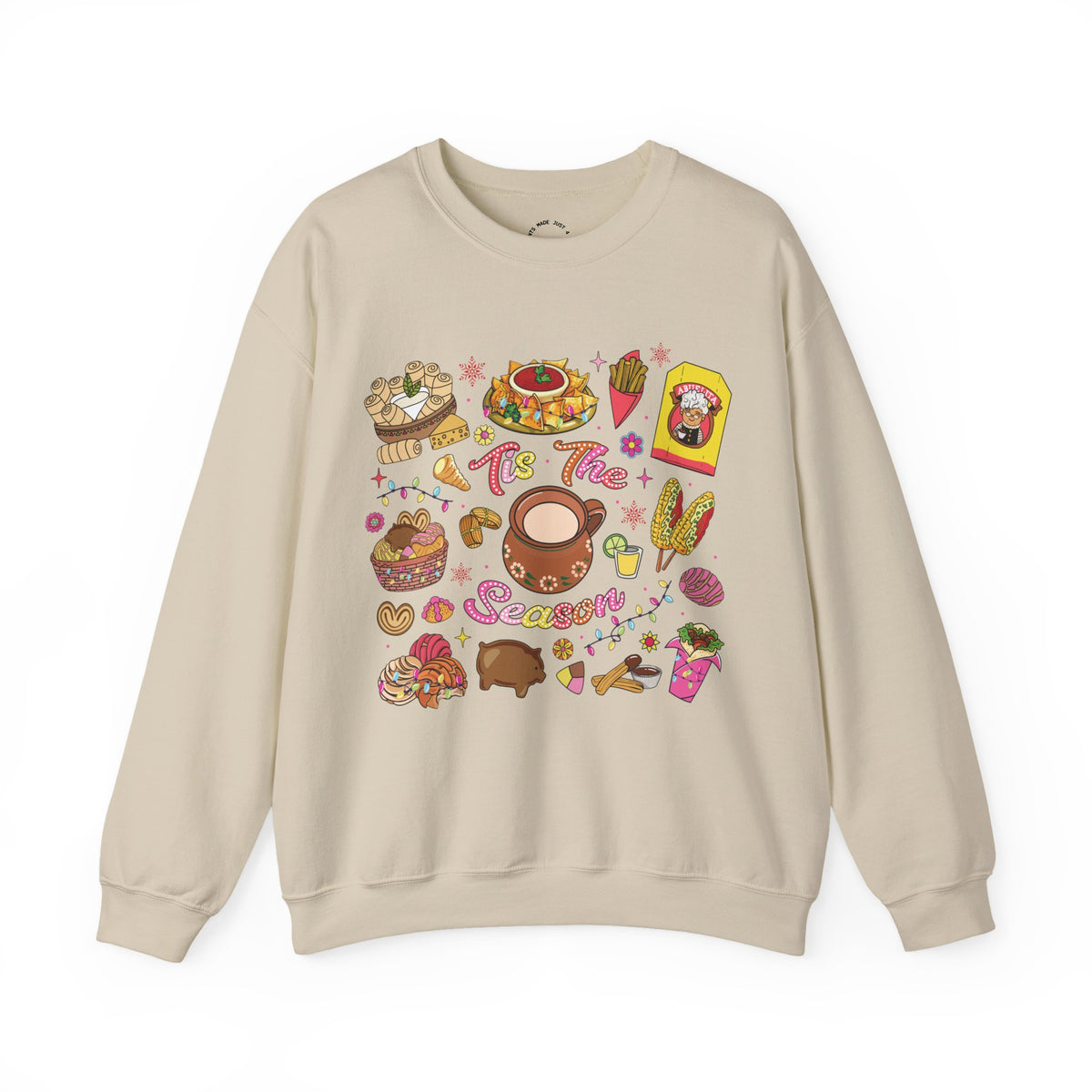 A beige crewneck sweatshirt featuring a colorful Christmas-themed graphic print with illustrations of seasonal hispanic treats and the text 'Tis The Season'.