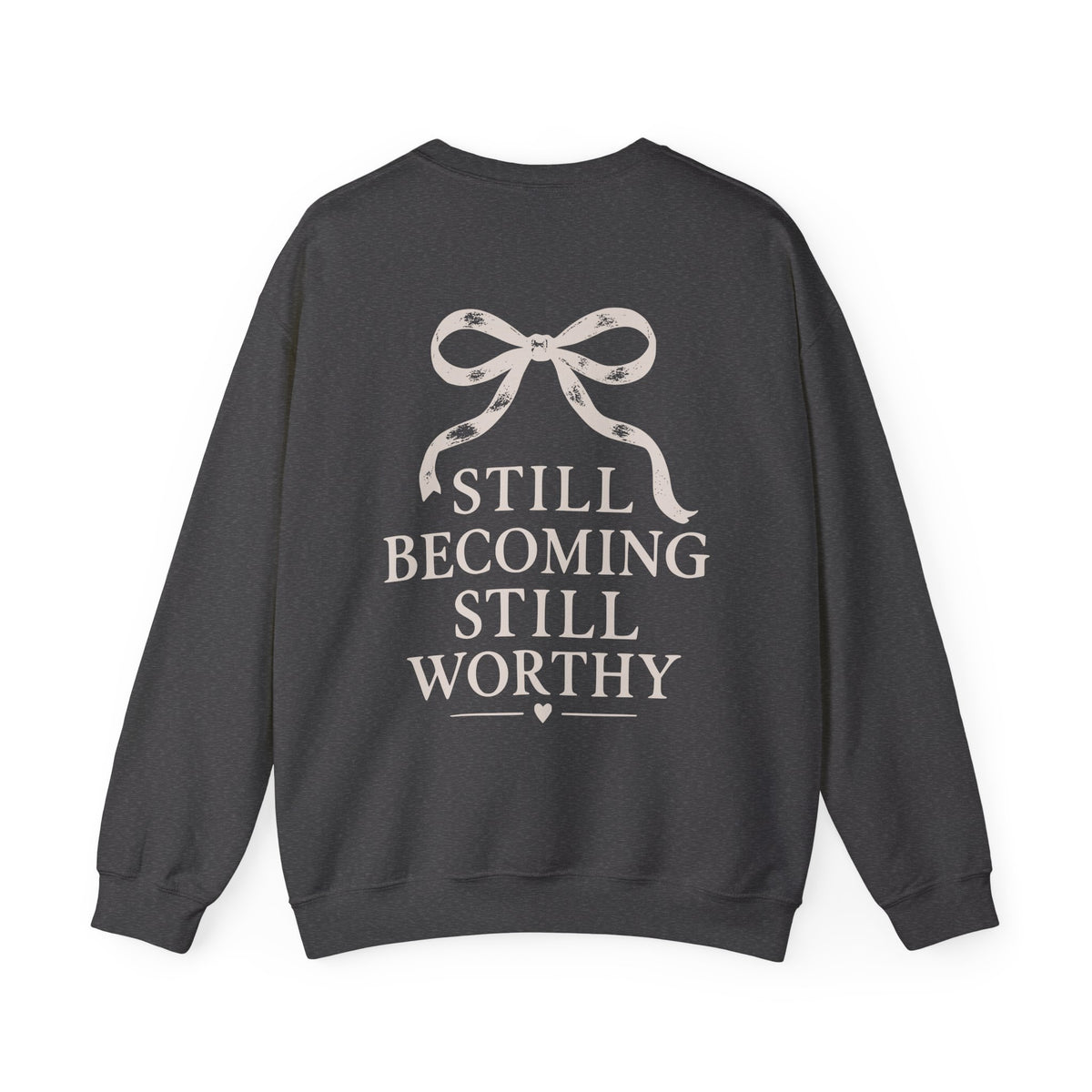 A dark charcoal gray crewneck sweatshirt shown from the back, featuring a cream-colored graphic of a ribbon bow above the text 'STILL BECOMING STILL WORTHY' in a serif font.