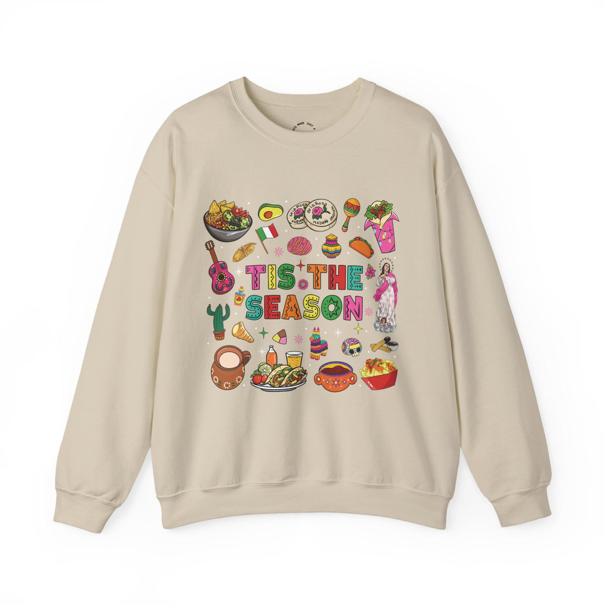 A beige crewneck sweatshirt featuring a colorful graphic print on the front with the text 'Tis the Season' surrounded by various cozy mexican illustrations like hot cocoa, burritos, tacos, pinata, and more seasonal traditions.