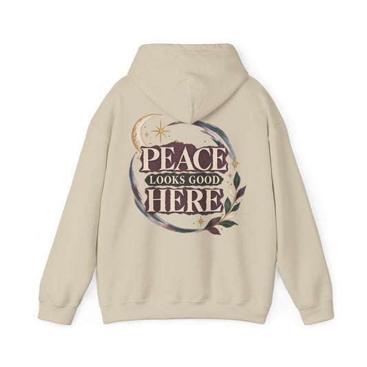 A beige hoodie shown from the back, featuring a large graphic print with the text 'PEACE LOOKS GOOD HERE' inside a circular wreath design with stars and leaves.