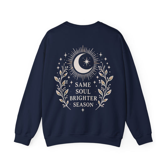 A navy blue crewneck sweatshirt shown from the back, featuring a cream graphic of a crescent moon, stars, and leafy branches with the text 'SAME SOUL BRIGHTER SEASON'.