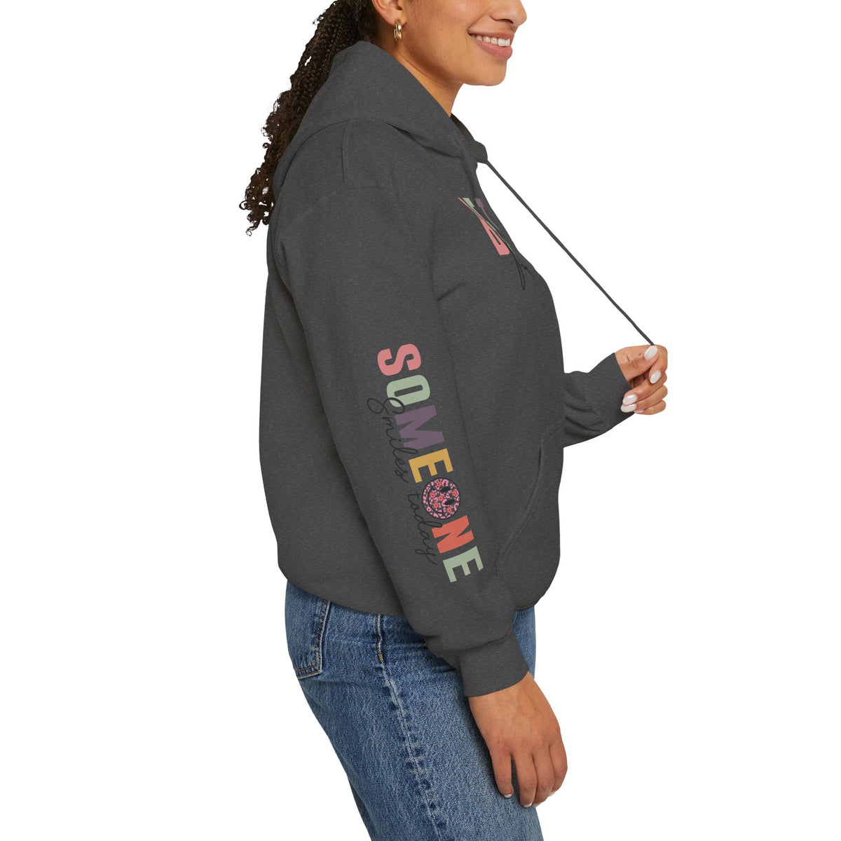 A woman wearing a dark gray heather hoodie with text graphics on the right sleeve 'Someone Smiles today' with a smiley face, paired with blue jeans, shown from a side profile against a white background.