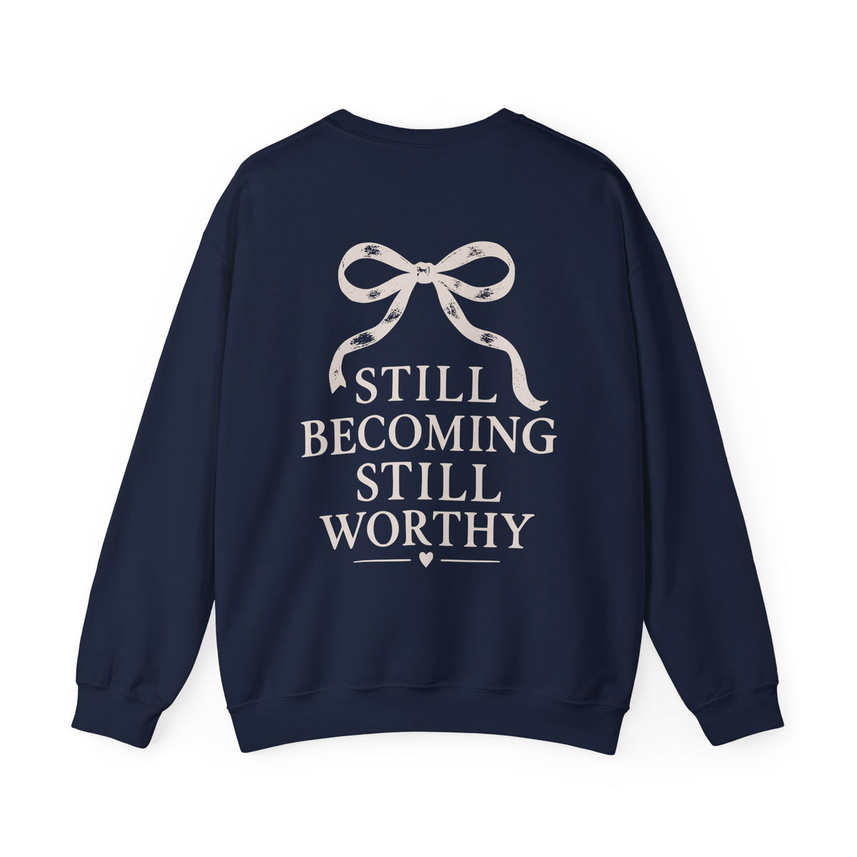 A navy blue crewneck sweatshirt from the back with a cream graphic featuring a ribbon bow and the text 'STILL BECOMING STILL WORTHY' in a serif font, shown against a plain white background.