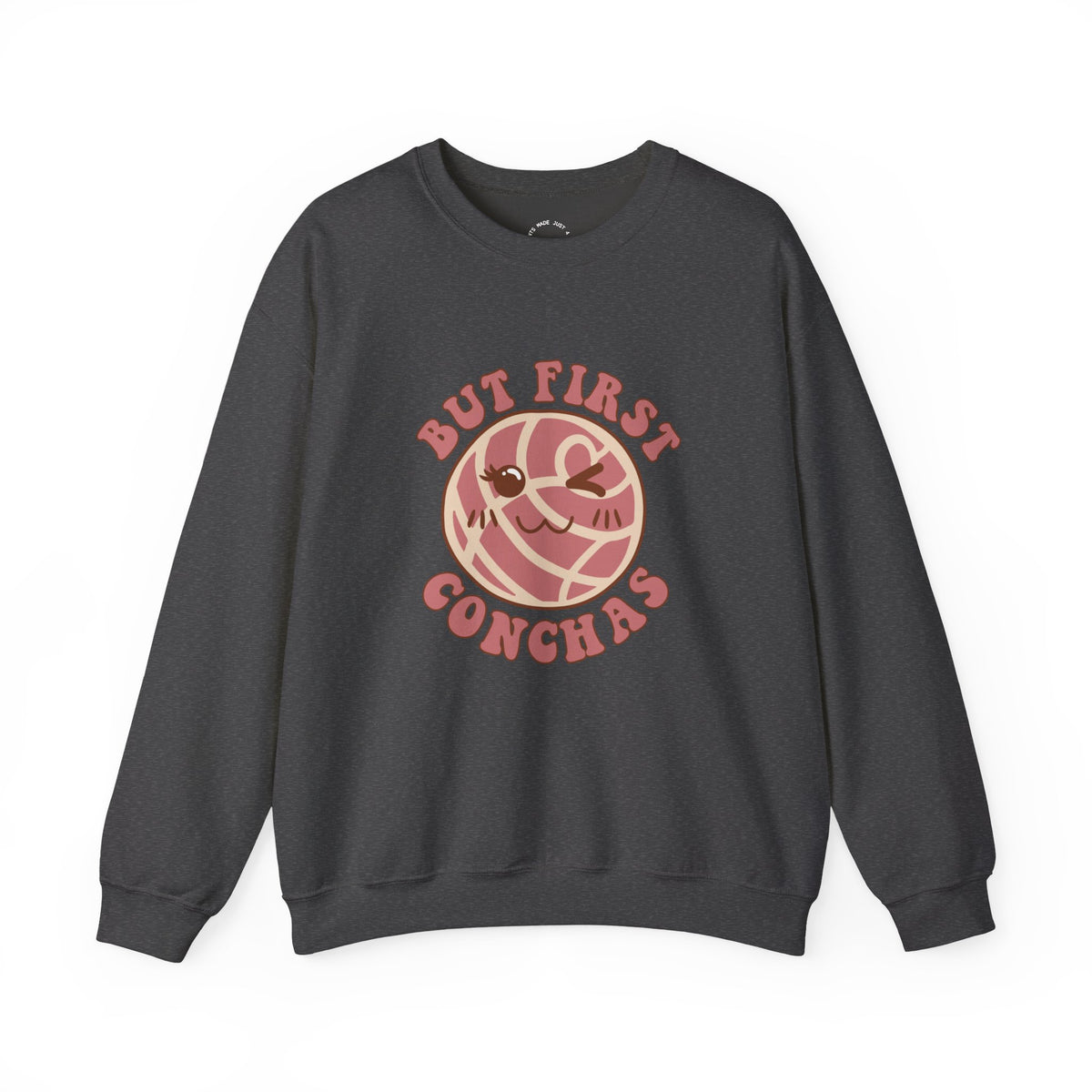 Dark Heather crewneck sweatshirt featuring a graphic of a winking concha with the text 'BUT FIRST CONCHAS' in pinkish-red lettering.