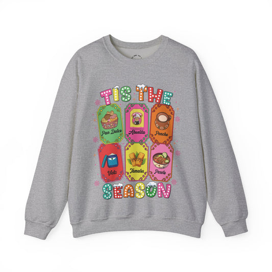 A heather gray crewneck sweatshirt featuring a colorful graphic print of six Christmas-themed ticket illustrations with Latino traditions with the text 'TIS THE SEASON' in a retro, bubbly font.