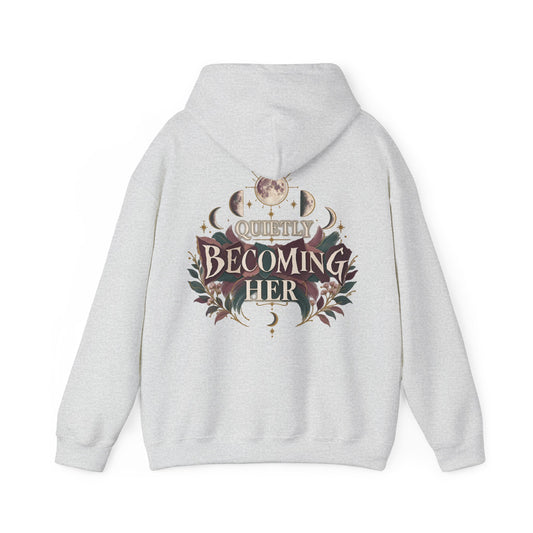 A light heather gray pullover hoodie shown from the back, featuring a large graphic print with the words 'Quietly Becoming Her' surrounded by decorative floral and celestial motifs in muted maroons, green, and gold tones.