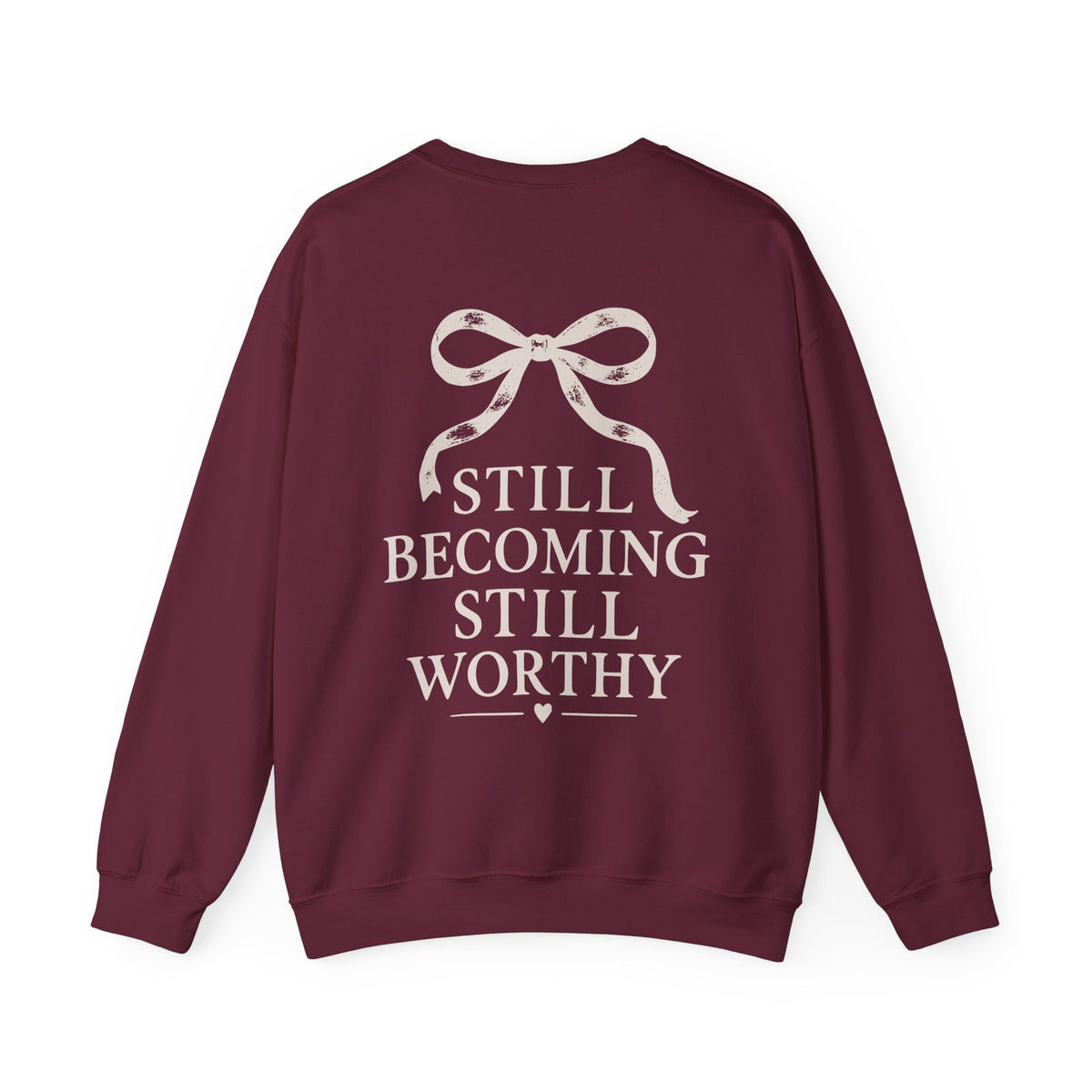 A maroon crewneck sweatshirt with a cream graphic on the back featuring a ribbon bow and the text 'STILL BECOMING STILL WORTHY'.