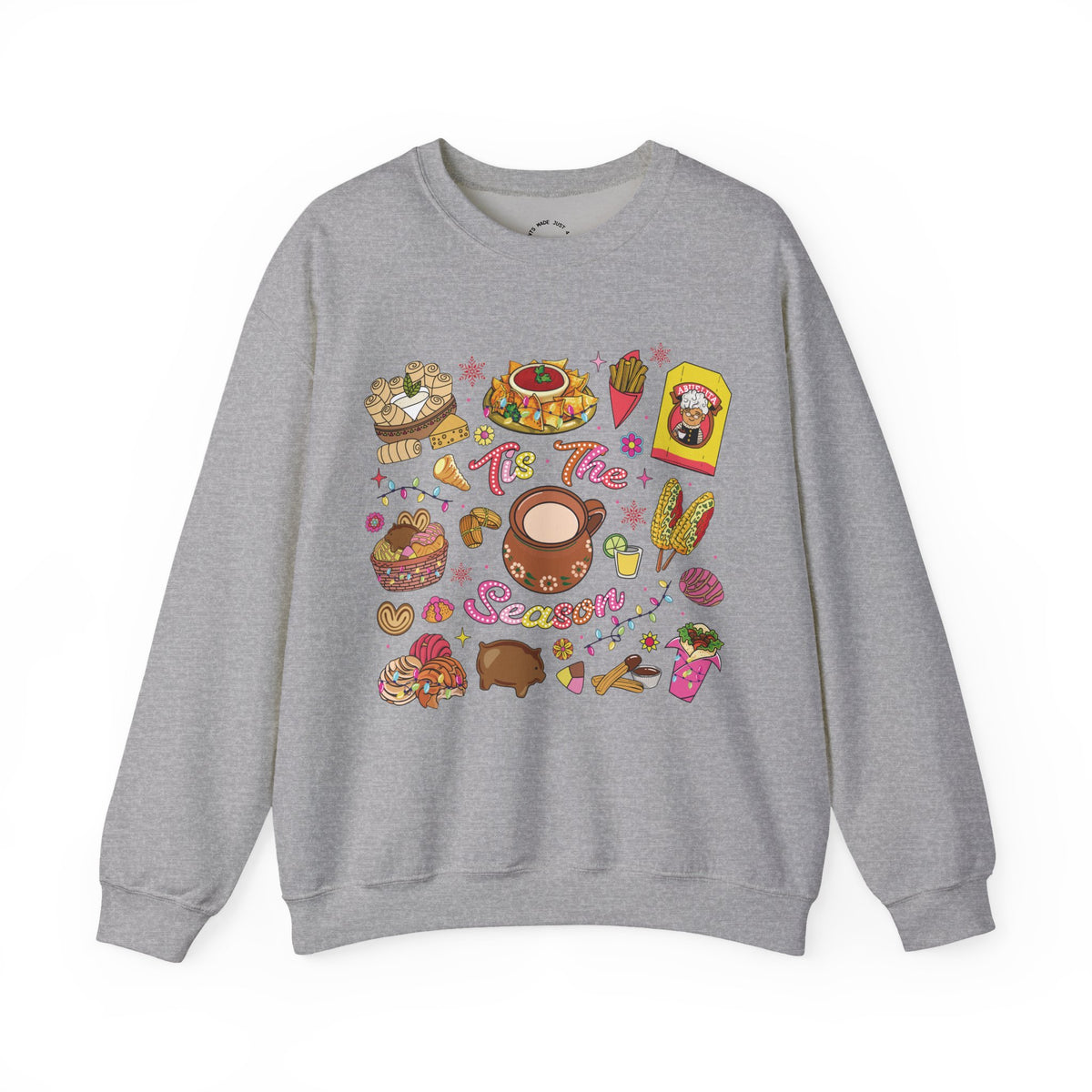 A heather gray crewneck sweatshirt featuring a colorful Christmas-themed graphic print with illustrations of seasonal hispanic treats and the text 'Tis The Season'.