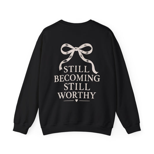 A black crewneck sweatshirt with a graphic on the back featuring a ribbon bow and the text 'STILL BECOMING STILL WORTHY' in a light beige color, shown against a plain white background.