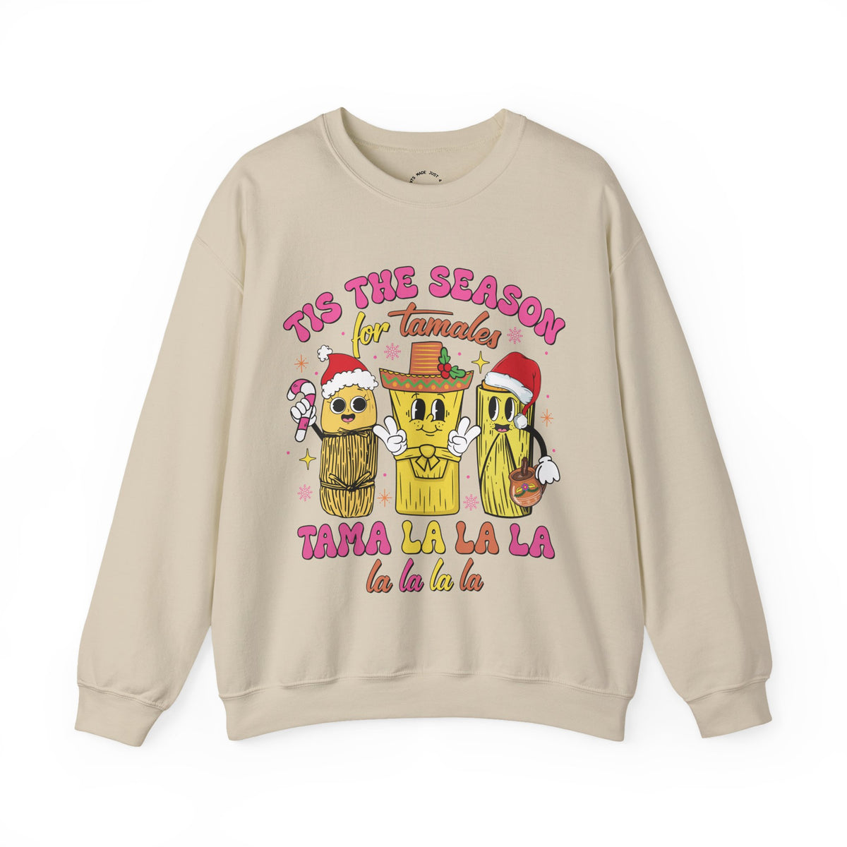 A beige crewneck sweatshirt featuring a colorful graphic of three tamale characters wearing Santa hats and a sombrero, with the text 'TIS THE SEASON for Tamales' and 'TAMA LA LA LA la la la la'.