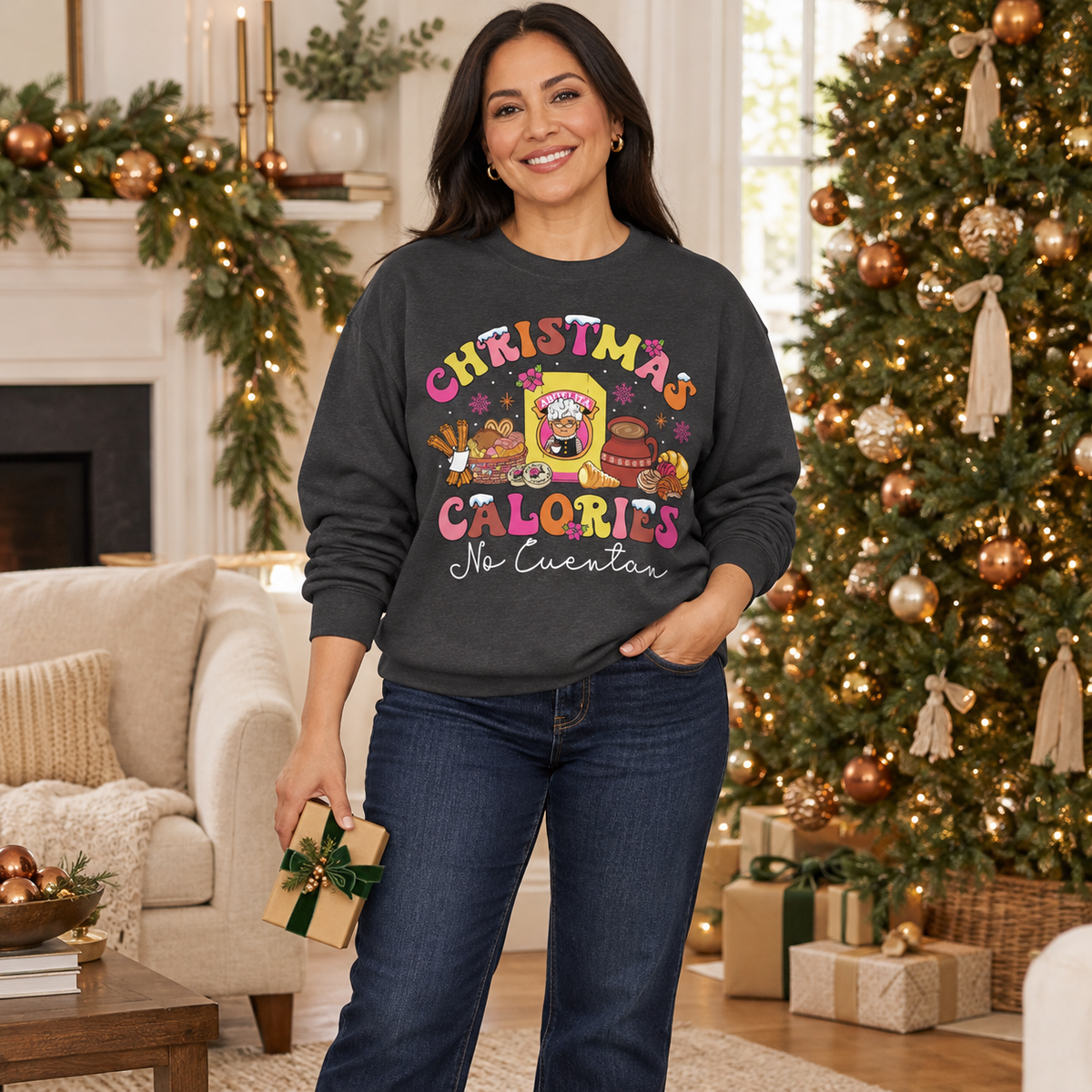 A woman stands in a cozy living room decorated for Christmas, wearing a dark charcoal gray sweatshirt with a colorful graphic that reads 'CHRISTMAS CALORIES No Cuentan' and blue jeans.