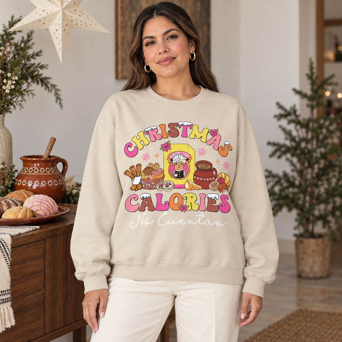 A woman wearing a beige crewneck sweatshirt with a colorful cartoon graphic that reads 'CHRISTMAS CALORIES No Cuentan' in white script, posed in a warm, decorated holiday room.