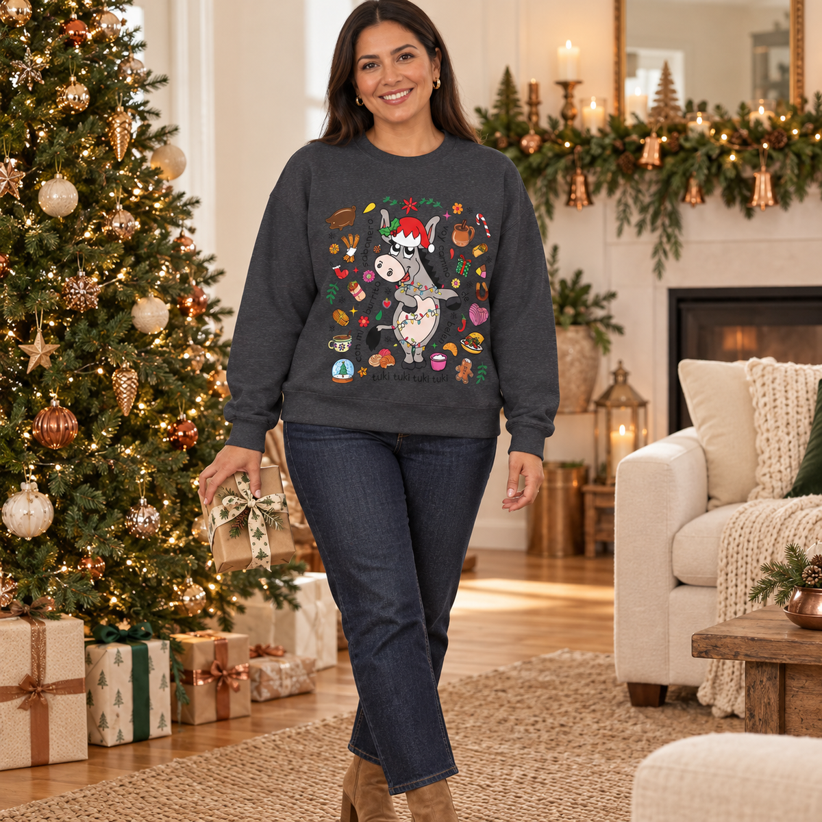 A woman wearing a dark gray crewneck sweatshirt with a colorful Christmas donkey graphic, paired with dark blue jeans, standing in a decorated living room next to a Christmas tree.