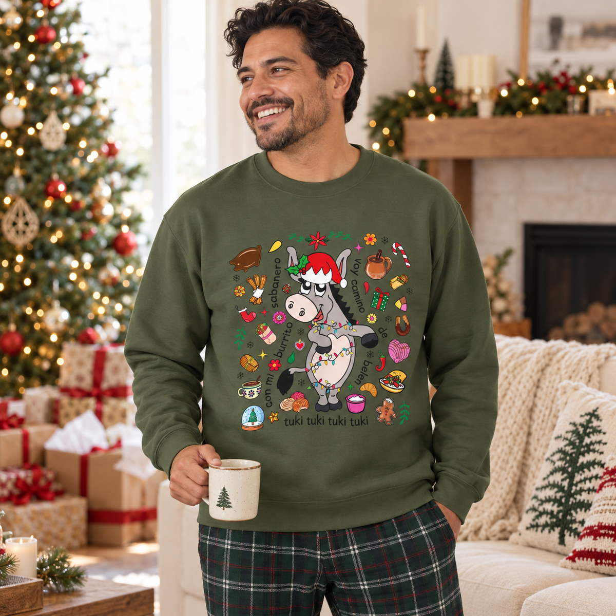 A man wearing an olive green crewneck sweatshirt with a festive Christmas donkey graphic, standing in a cozy living room with a decorated Christmas tree and fireplace in the background.