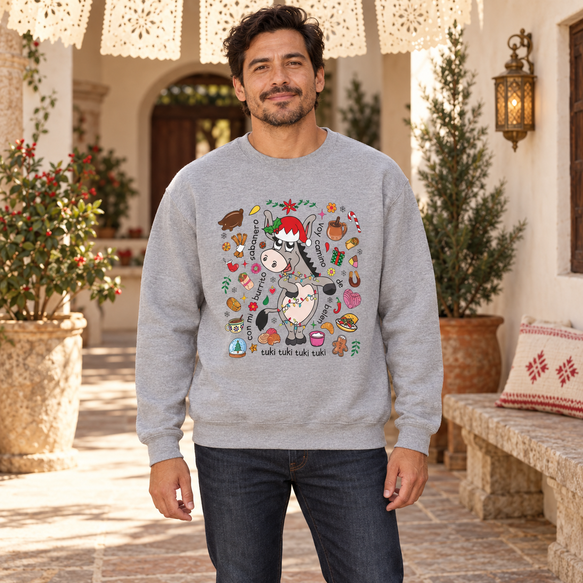 A man wearing a light grey crewneck sweatshirt with a colorful cartoon donkey graphic and Spanish text 'tuki tuki tuki tuki' on the front, standing in a cozy, decorated patio setting.