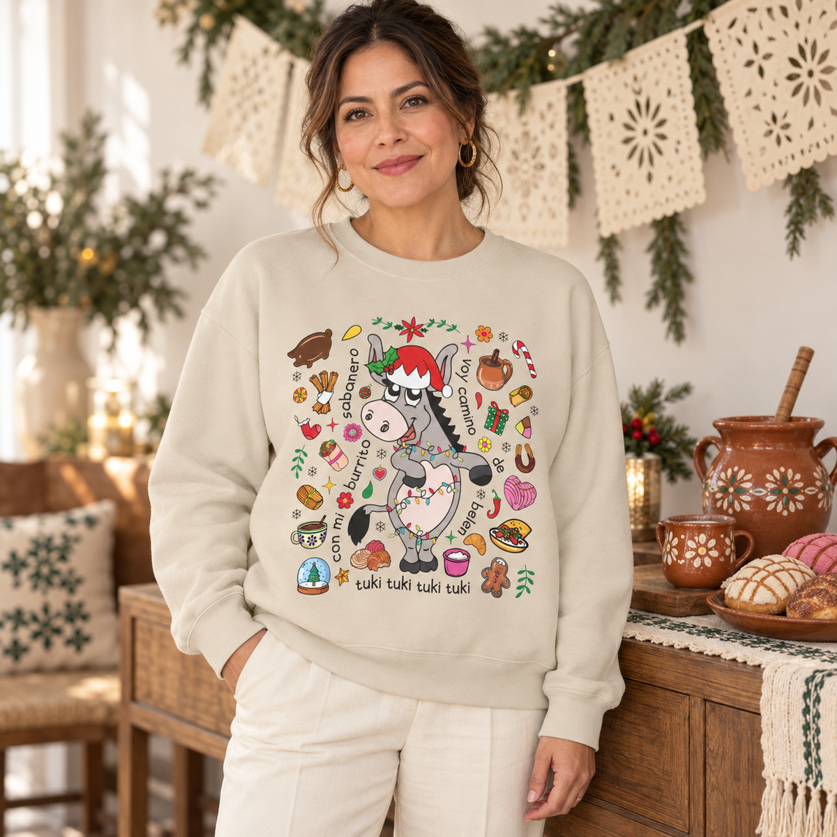 A woman wearing a cream-colored crewneck sweatshirt with a colorful cartoon donkey graphic and Spanish text 'tuki tuki tuki tuki' on the front, standing in a cozy, decorated kitchen setting.