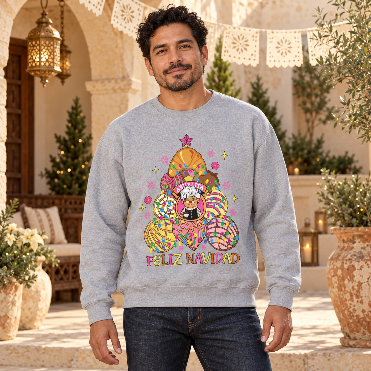 A man wearing a light gray crewneck sweatshirt with a colorful concha christmas tree with abuelita in the middle and the text 'FELIZ NAVIDAD' under the tree. The background is a warm, festive outdoor patio with arches, lanterns, and potted plants.