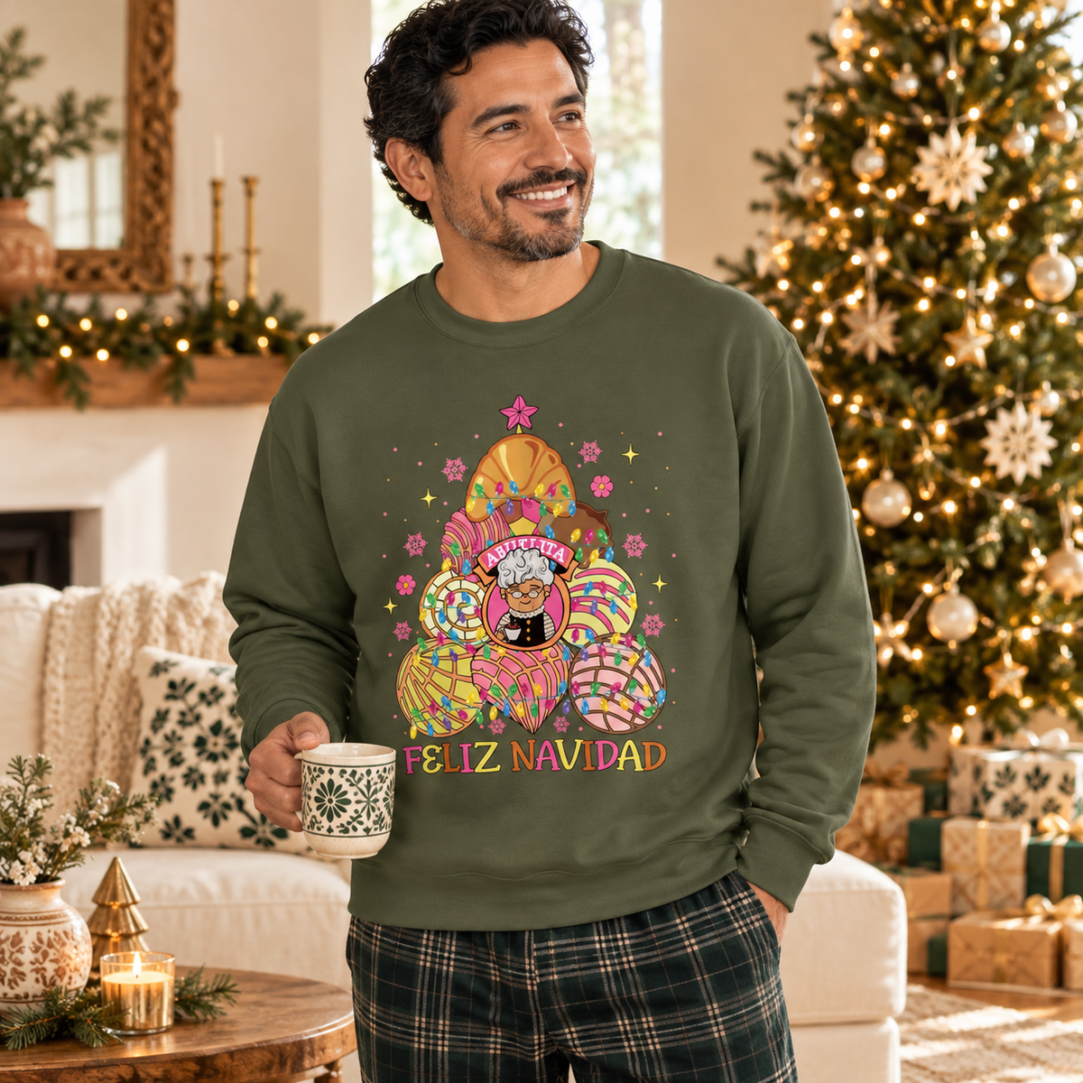 A man wearing an olive green crewneck sweatshirt with a colorful festive graphic of colorful concha christmas tree with abuelita in the middle and the text 'FELIZ NAVIDAD' under the tree, holding a mug in a cozy living room decorated for Christmas.