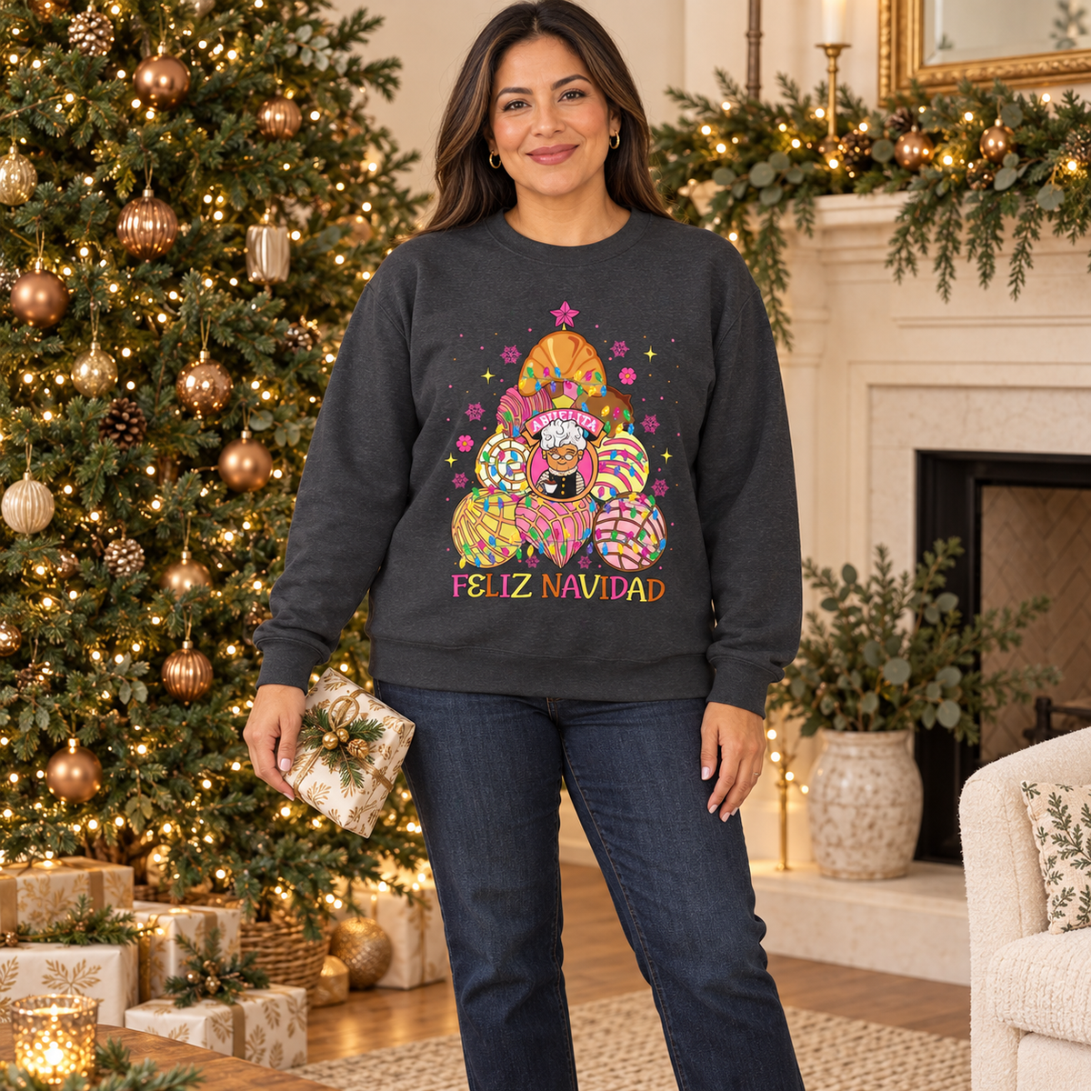A woman wearing a dark charcoal gray crewneck sweatshirt with colorful concha christmas tree with abuelita in the middle and the text 'FELIZ NAVIDAD' under the tree, she stands in a decorated living room in front of a Christmas tree and fireplace.