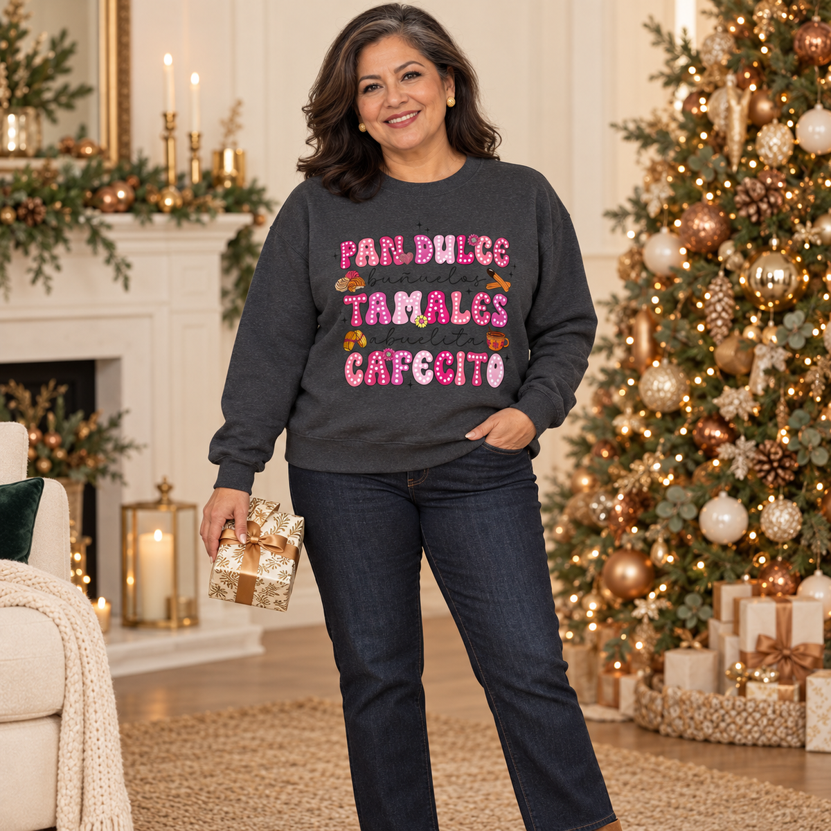 A woman stands in a decorated holiday living room wearing a dark gray sweatshirt with print on the front with the Spanish text 'Pan Dulce Bunuelas Tamales Abuelita Cafecito' in pink and brown donut-style lettering with small illustrations of food items. and dark blue jeans. She is holding a small wrapped gift box, with a Christmas tree, fireplace, and candles in the background.