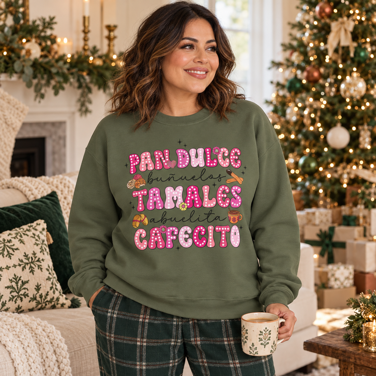 A woman wearing an olive green crewneck sweatshirt with a print on the front with the Spanish text 'Pan Dulce Bunuelas Tamales Abuelita Cafecito' in pink and brown donut-style lettering with small illustrations of food items in a decorated Christmas living room setting.