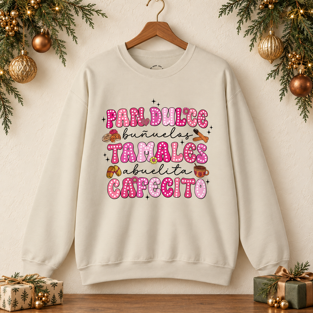 A beige crewneck sweatshirt hanging on a wooden hanger against a warm beige wall, decorated with Christmas greenery, gold ornaments, and wrapped gifts on a wooden surface. The sweatshirt features print on the front with the Spanish text 'Pan Dulce Bunuelas Tamales Abuelita Cafecito' in pink and brown donut-style lettering with small illustrations of food items.