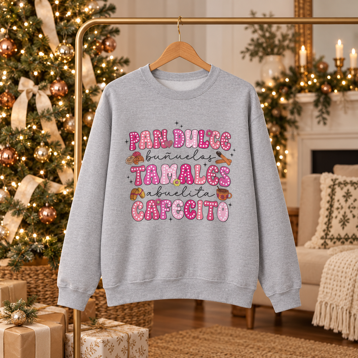 A light gray crewneck sweatshirt hanging on a wooden hanger from a gold clothing rack, featuring a print on the front with the Spanish text 'Pan Dulce Bunuelas Tamales Abuelita Cafecito' in pink and brown donut-style lettering with small illustrations of food items. The background depicts a cozy, warmly lit Christmas living room with a decorated tree, fireplace, and sofa.