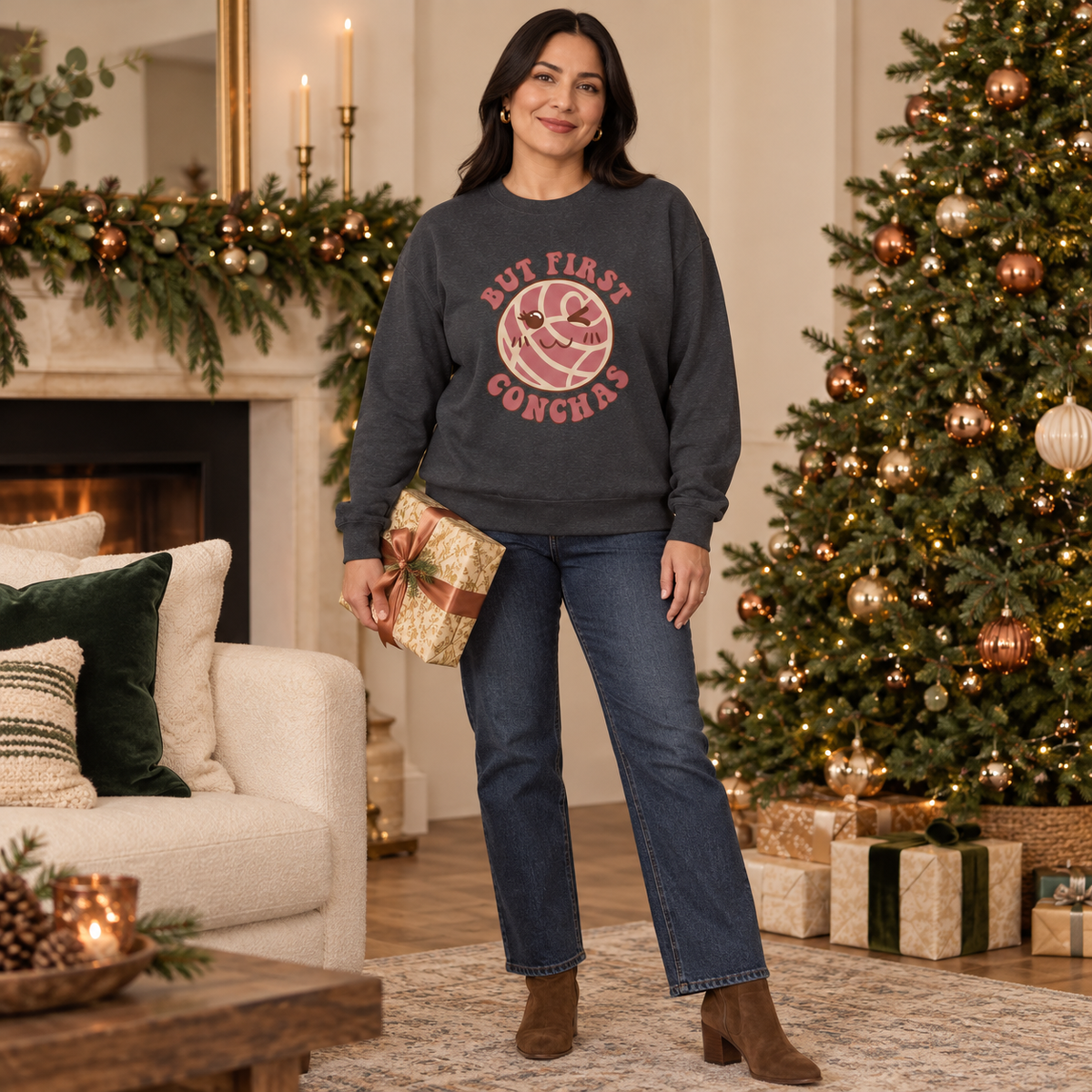 A woman stands in a decorated holiday living room wearing a dark charcoal crewneck sweatshirt with a pink graphic that reads 'BUT FIRST CONCHAS'. She is also wearing blue jeans and brown boots, holding a wrapped gift box.