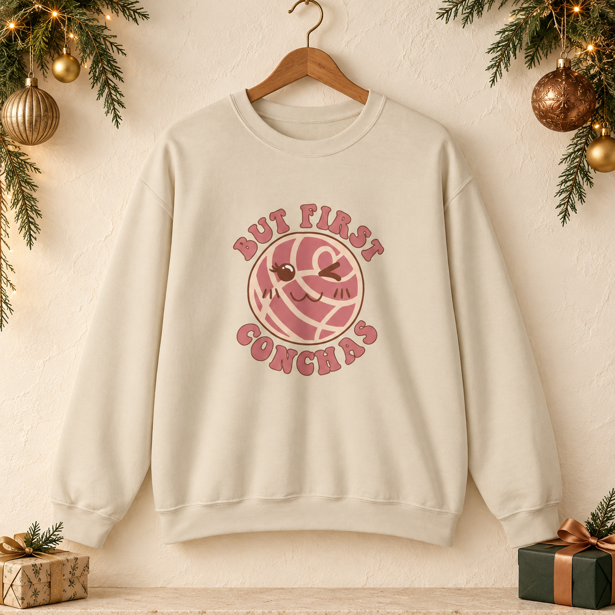 Beige crewneck sweatshirt featuring a graphic of a winking concha with the text 'BUT FIRST CONCHAS' in pinkish-red lettering.