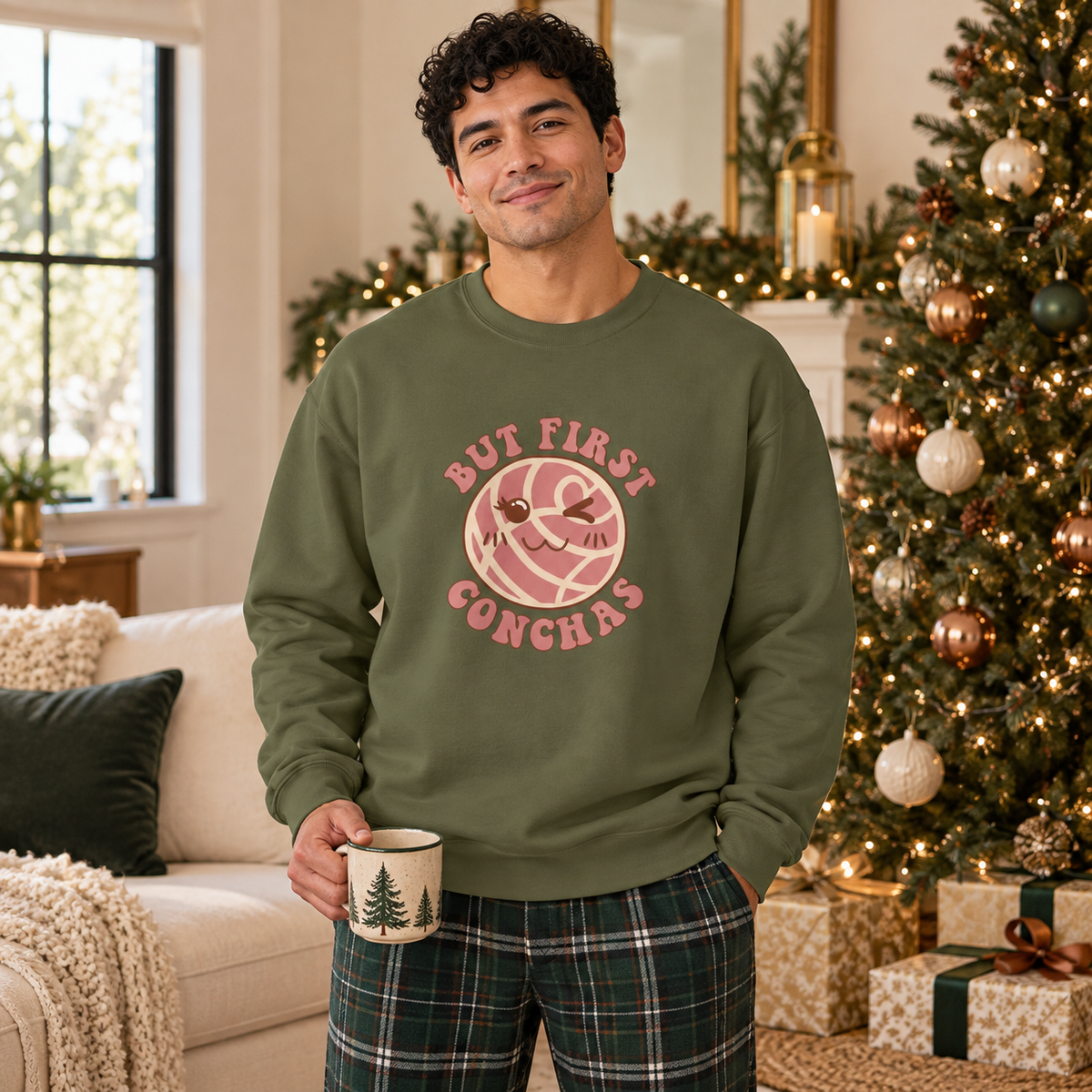 A man wearing an olive green crewneck sweatshirt with a pink graphic that reads 'BUT FIRST CONCHAS' and a plaid pajama pants, holding a mug in a cozy, decorated living room with a Christmas tree.