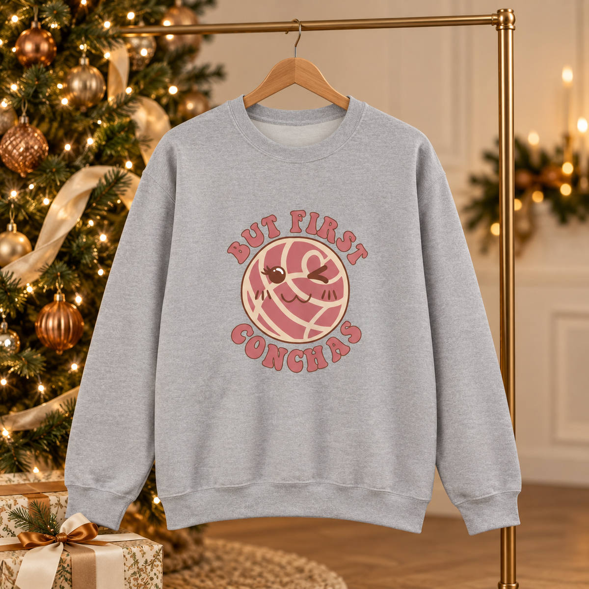 A light heather gray crewneck sweatshirt hanging on a wooden hanger from a gold clothing rack, set against a warm, festive Christmas tree background. The sweatshirt features a graphic of a pink and white concha with a small smiling face, surrounded by the text 'BUT FIRST CONCHAS' in pink.