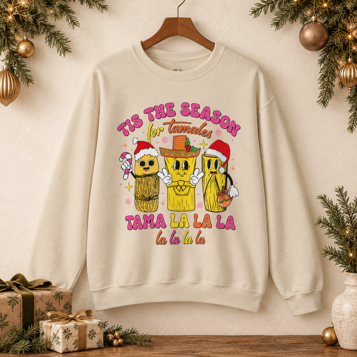 An beige crewneck sweatshirt hanging on a wooden hanger against a warm beige wall, decorated with Christmas greenery and ornaments. The sweatshirt features a colorful graphic of three tamale characters wearing Santa hats and a sombrero, with the text 'TIS THE SEASON for Tamales' and 'TAMA LA LA LA la la la la'.