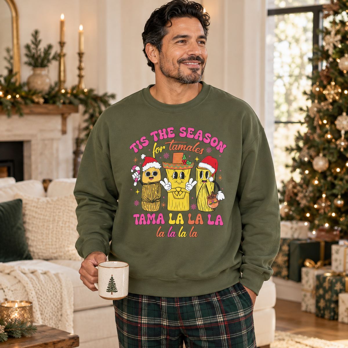 A man wearing an olive green crewneck sweatshirt with a colorful graphic of three tamale characters wearing Santa hats and a sombrero, with the text 'TIS THE SEASON for Tamales' and 'TAMA LA LA LA la la la la'. The background shows a cozy, decorated living room with a fireplace, holiday lights, and a Christmas tree.