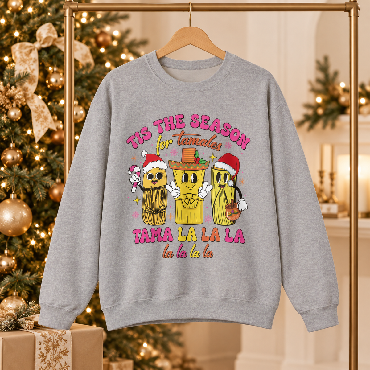 A light gray crewneck sweatshirt hanging on a wooden hanger, featuring a colorful graphic of three tamale characters wearing Santa hats and a sombrero, with the text 'TIS THE SEASON for Tamales' and 'TAMA LA LA LA la la la la'. The background shows a decorated Christmas tree and warm holiday lights.