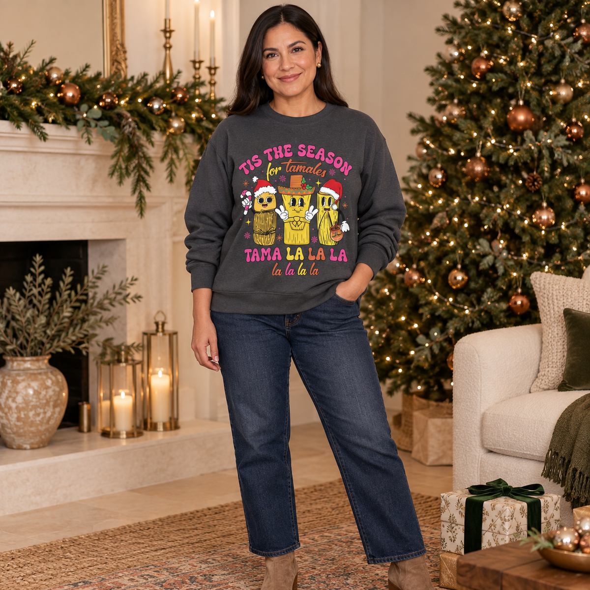 A woman stands in a decorated living room wearing a dark gray crewneck sweatshirt with a colorful Christmas graphic of three tamale characters wearing Santa hats and a sombrero, with the text 'TIS THE SEASON for Tamales' and 'TAMA LA LA LA la la la la'. She is also wearing dark blue jeans.