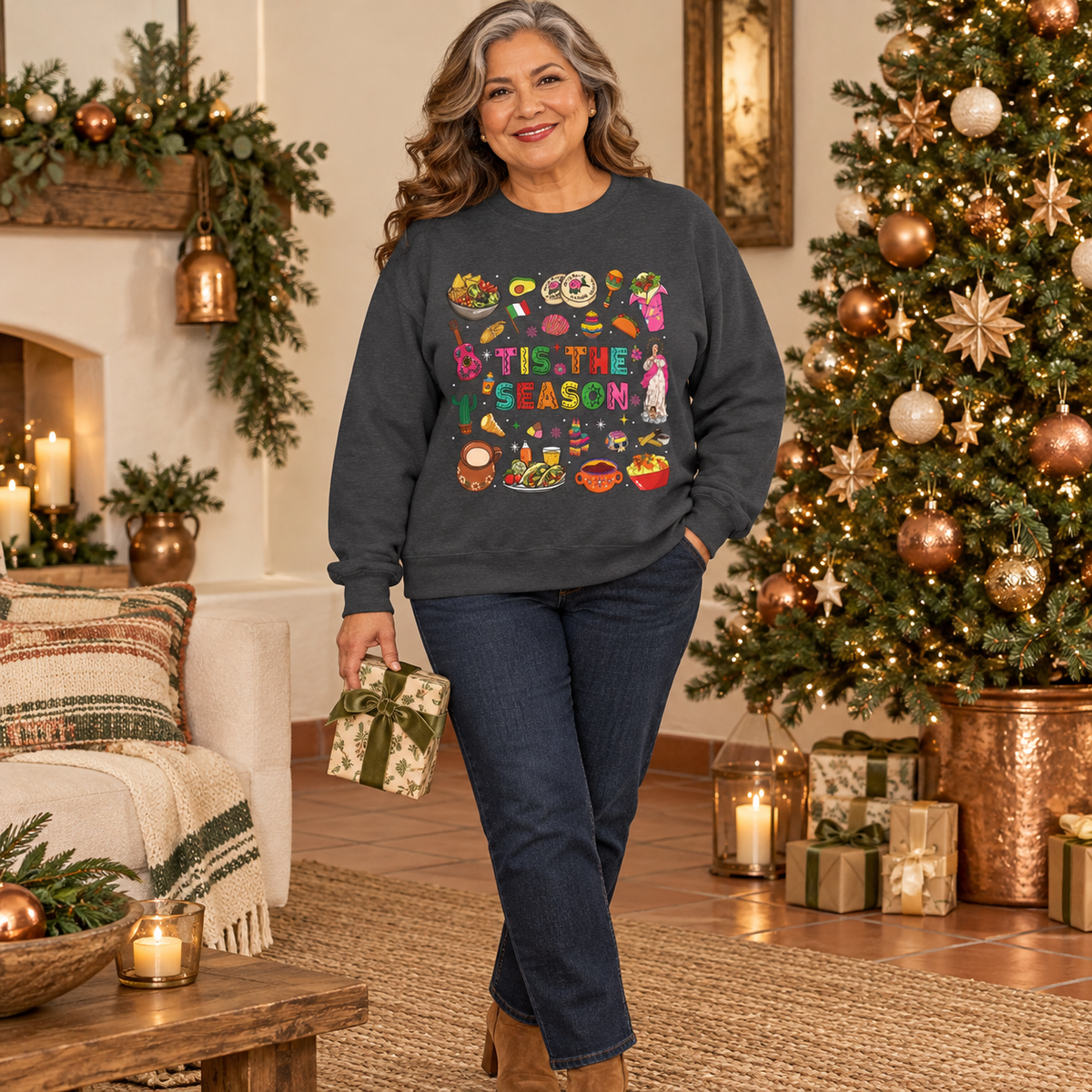 A woman stands in a decorated holiday living room wearing a dark gray crewneck sweatshirt featuring a colorful graphic print on the front with the text 'Tis the Season' surrounded by various cozy mexican illustrations like hot cocoa, burritos, tacos, pinata, and more seasonal traditions, paired with dark blue jeans.