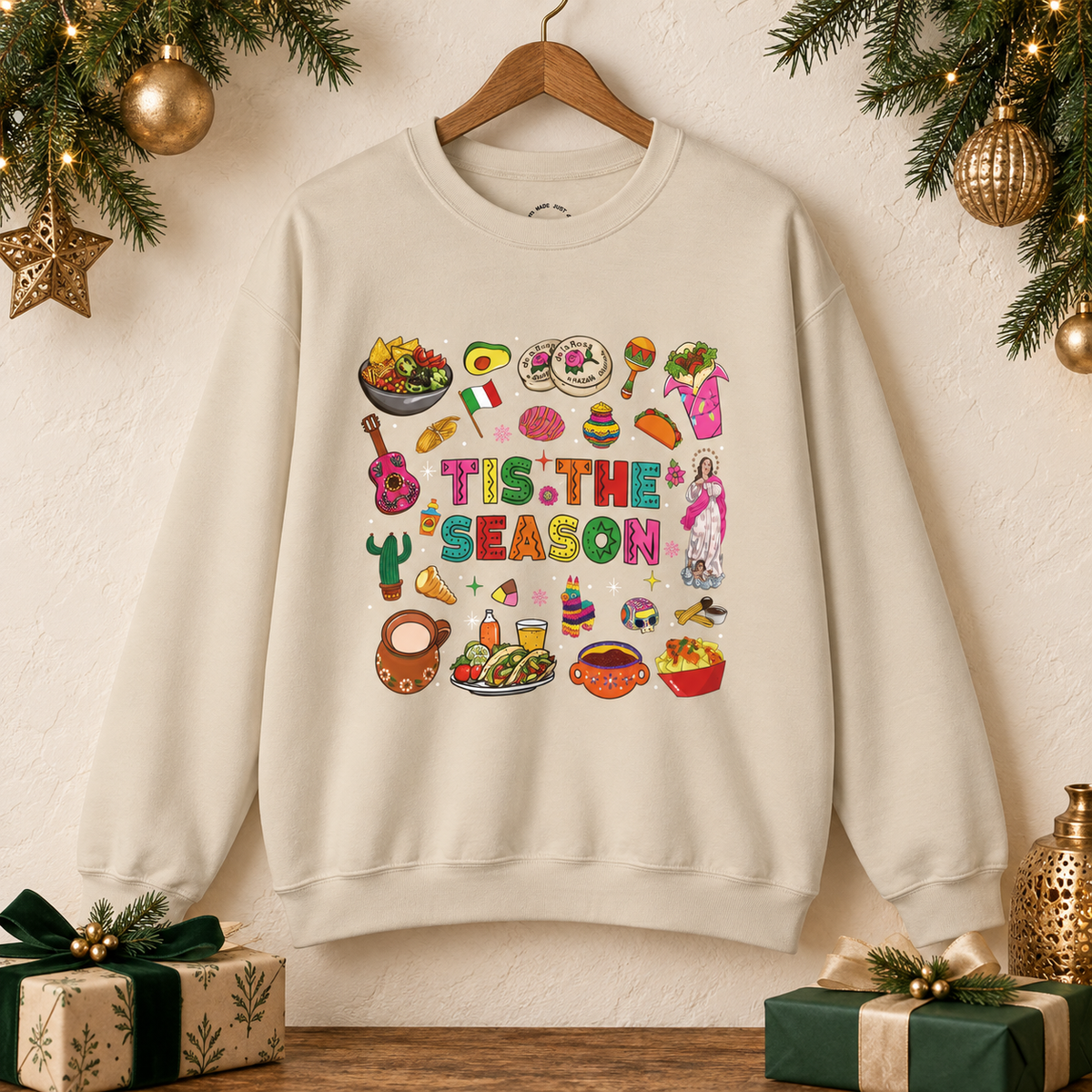 A beige crewneck sweatshirt hanging on a wooden hanger against a textured cream wall, decorated with Christmas greenery, gold ornaments, and wrapped gifts. The sweatshirt features a colorful graphic print on the front with the text 'Tis the Season' surrounded by various cozy mexican illustrations like hot cocoa, burritos, tacos, pinata, and more seasonal traditions.