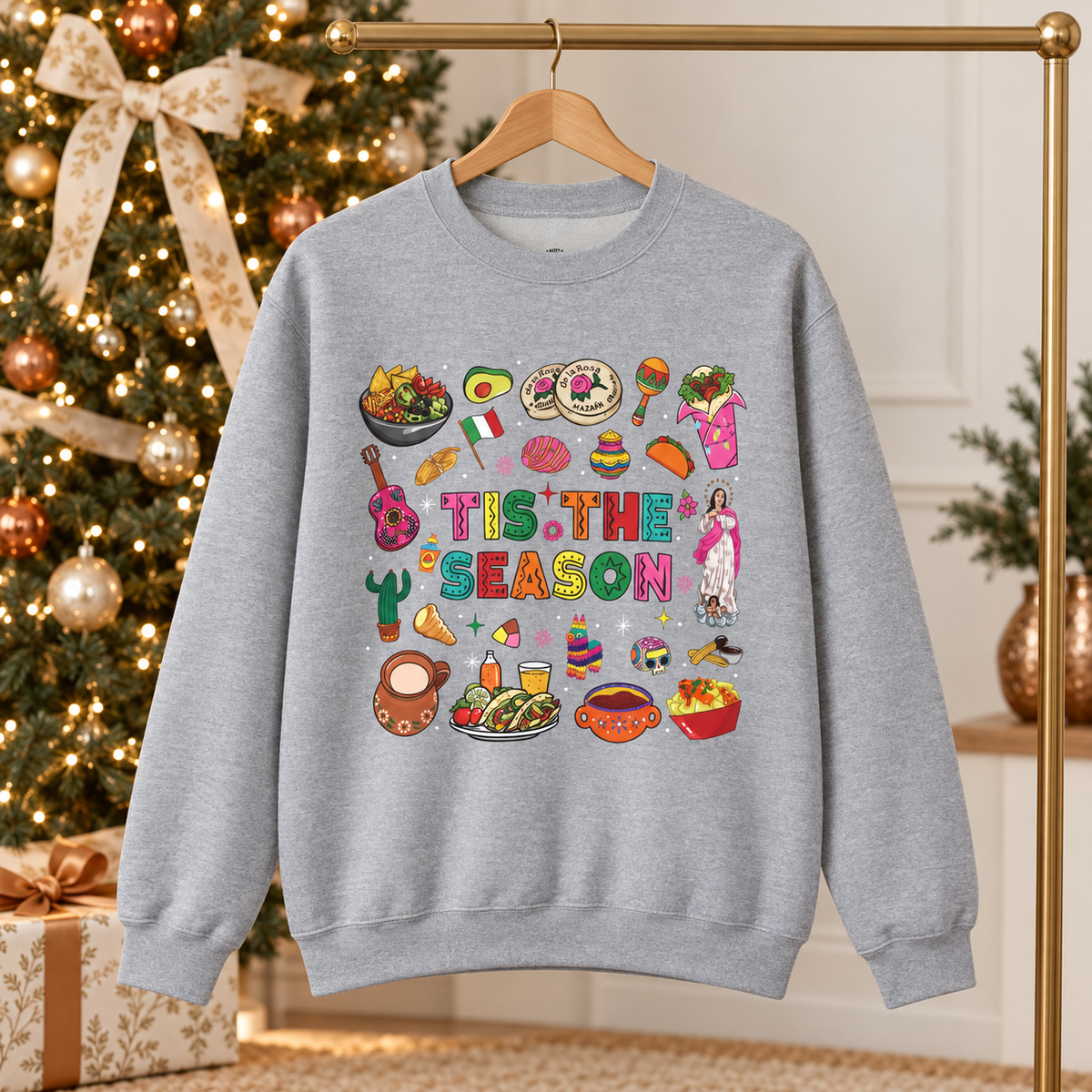 A light heather gray crewneck sweatshirt featuring a colorful graphic print on the front with the text 'Tis the Season' surrounded by various cozy mexican illustrations like hot cocoa, burritos, tacos, pinata, and more seasonal traditions. The background shows a decorated Christmas tree and holiday ornaments.