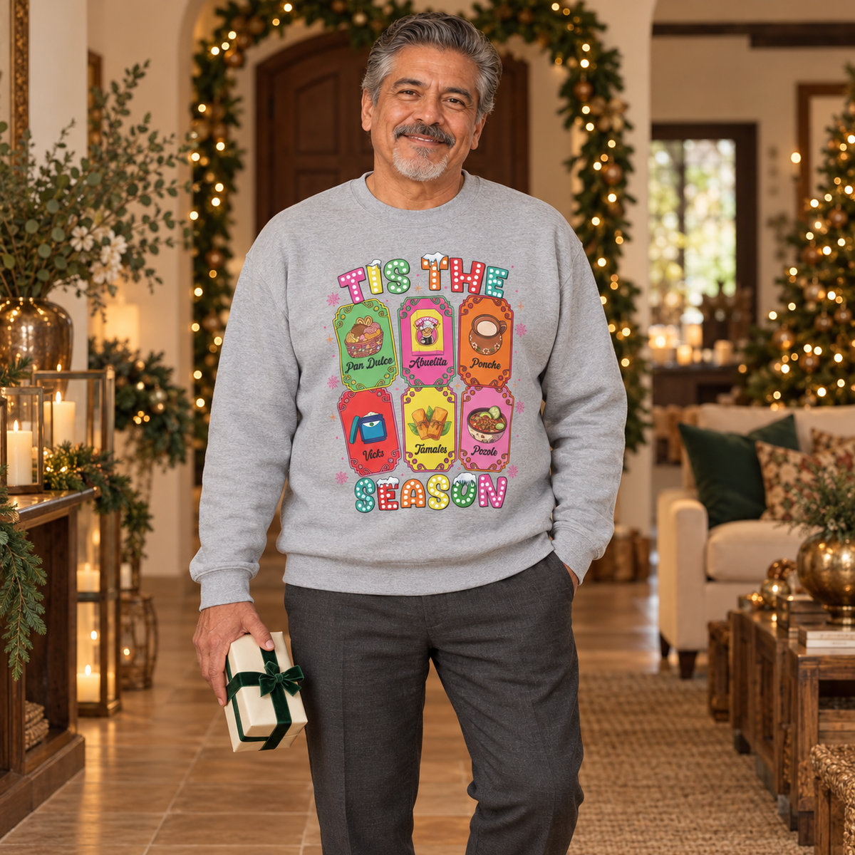 A man wearing a light gray crewneck sweatshirt featuring a colorful graphic print of six Christmas-themed ticket illustrations with Latino traditions with the text 'TIS THE SEASON' in a retro, bubbly font, standing in a warmly lit, decorated Christmas living room and holding a small gift box.