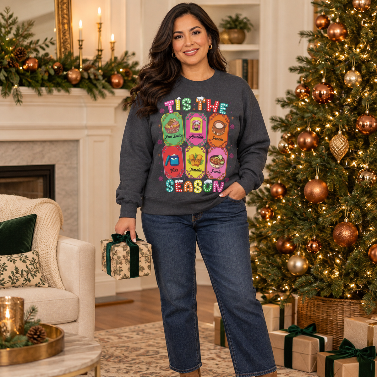 A woman stands in a decorated living room wearing a dark gray crewneck sweatshirt featuring a colorful graphic print of six Christmas-themed ticket illustrations with Latino traditions with the text 'TIS THE SEASON' in a retro, bubbly font and blue jeans, holding a wrapped gift box next to a Christmas tree and fireplace.