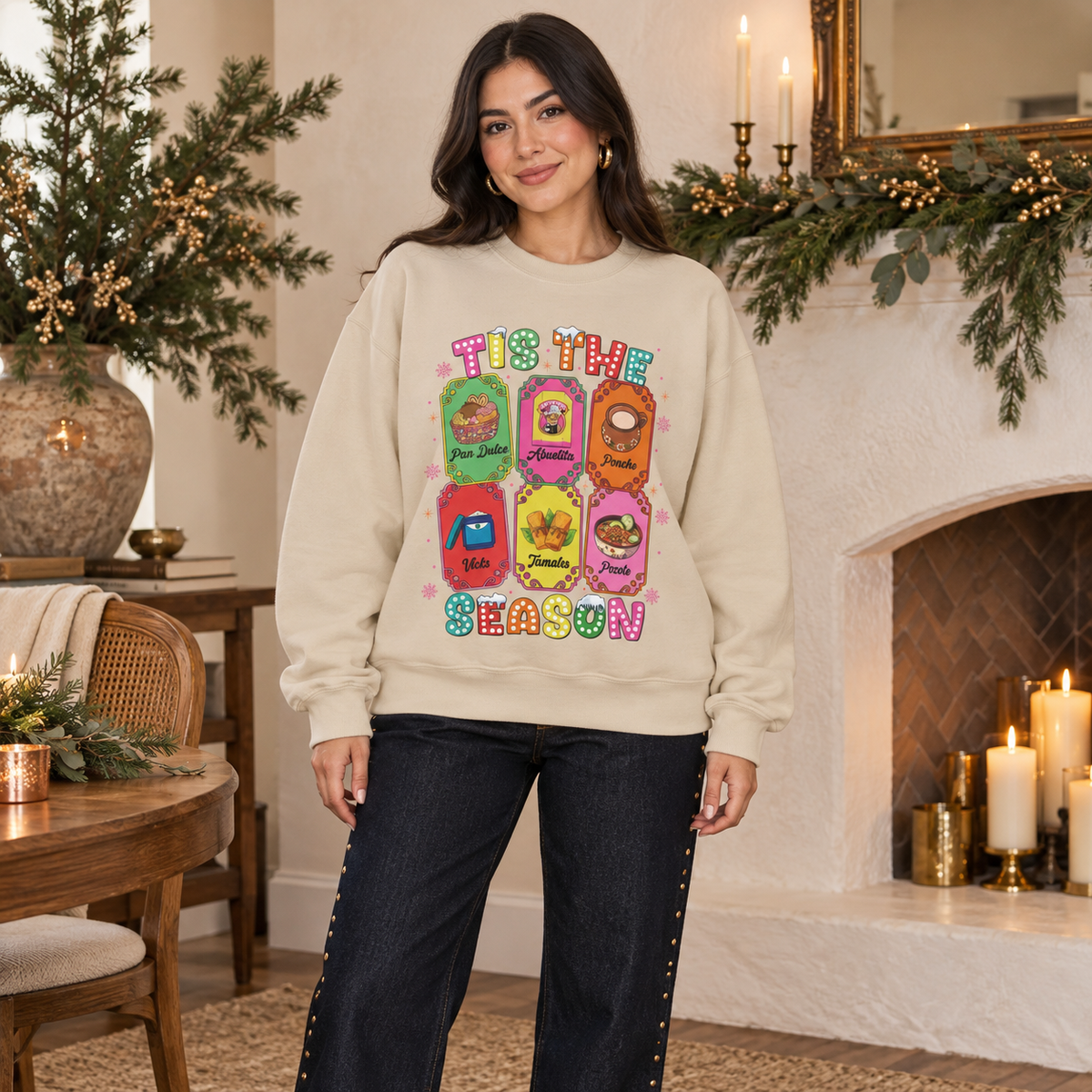 A woman stands indoors in front of a fireplace wearing a beige crewneck sweatshirt featuring a colorful graphic print of six Christmas-themed ticket illustrations with Latino traditions with the text 'TIS THE SEASON' in a retro, bubbly font and dark jeans.