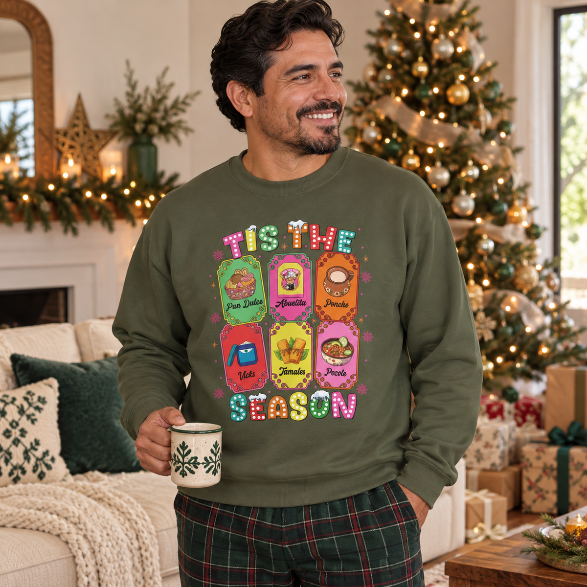 A man wearing an olive green crewneck sweatshirt featuring a colorful graphic print of six Christmas-themed ticket illustrations with Latino traditions with the text 'TIS THE SEASON' in a retro, bubbly font, standing in a cozy living room decorated for Christmas and holding a mug.